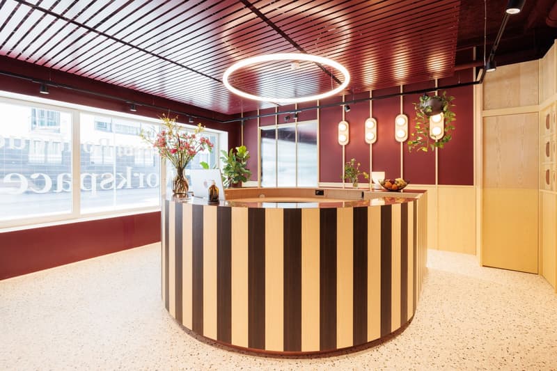 Spacious reception area with a circular wooden reception desk, modern lighting fixtures, and decorative plants. The walls are painted in a deep red color, creating a warm and inviting atmosphere. Large windows provide natural light and a view of the exter
