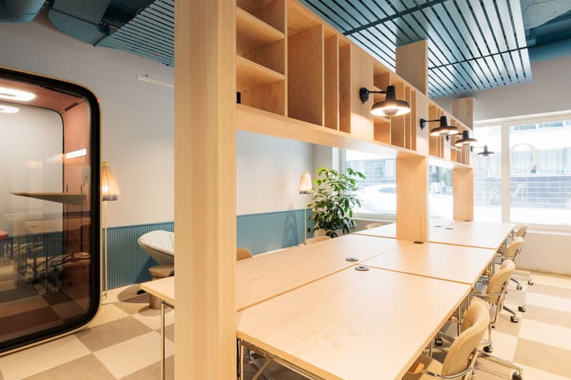 The image shows a modern and open office space with a large wooden conference table, shelving units along the walls, and various lighting fixtures. The space has a minimalist and clean design aesthetic, with a mix of natural wood tones and neutral colors.