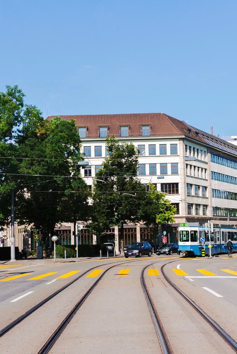 The image shows a street scene with a multi-story building, trees, and a tram track running through the middle. There are parked cars visible on the street, indicating the presence of parking spaces. The building has a traditional architectural style with