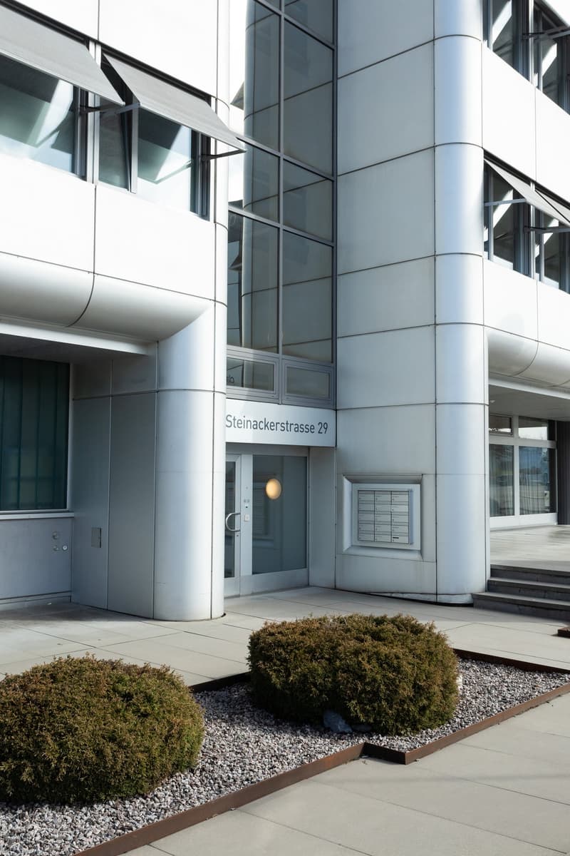 The image shows the exterior of a modern, multi-story building with a glass and metal facade. The building has a prominent address sign that reads 'Steinackerstrasse 29'. There are several large windows and glass panels on the facade, as well as some land