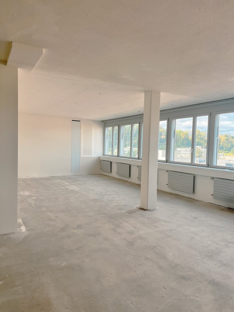 Large open-plan office space with white walls, concrete floor, and large windows providing natural light. The space appears to be in the process of renovation or construction, with visible heating units along the walls.