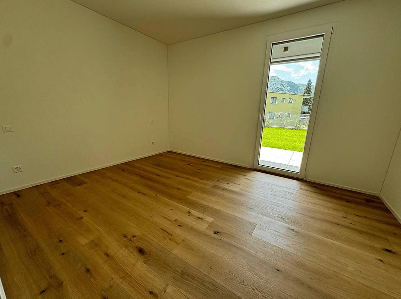 Spacious bedroom with hardwood flooring, neutral wall color, and a large window providing a view of the outdoor greenery.