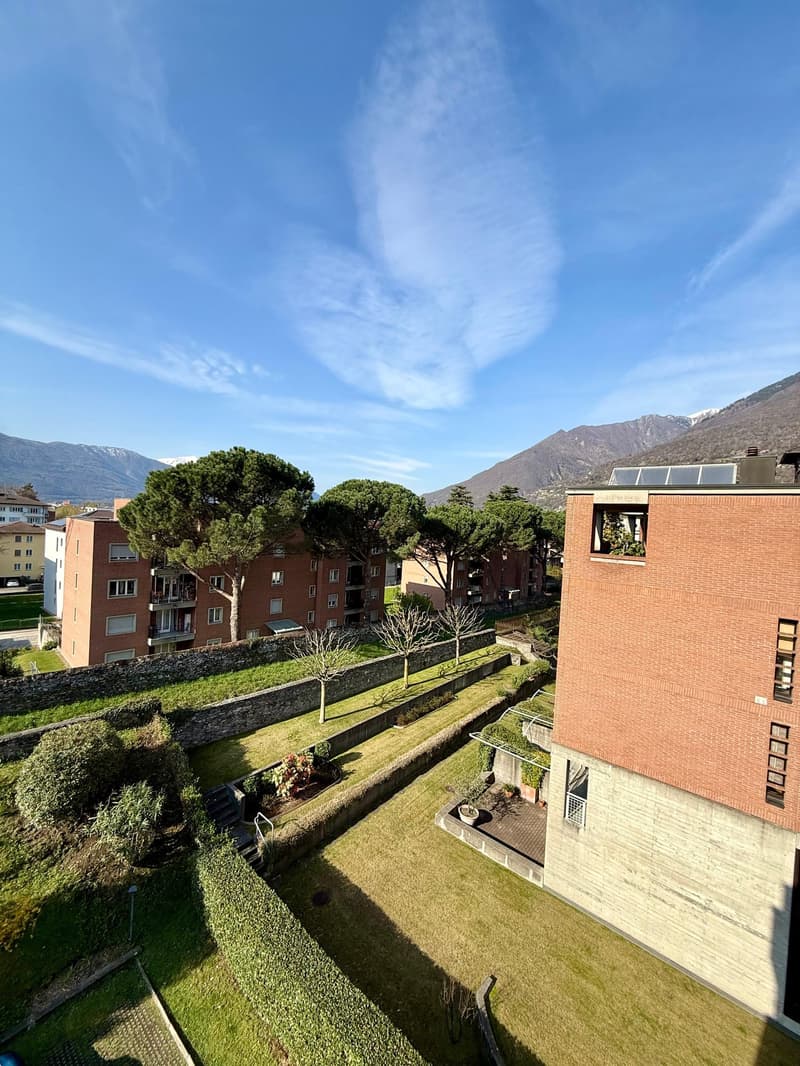 Aerial view of a building complex with multiple buildings, surrounded by greenery and mountains in the background.