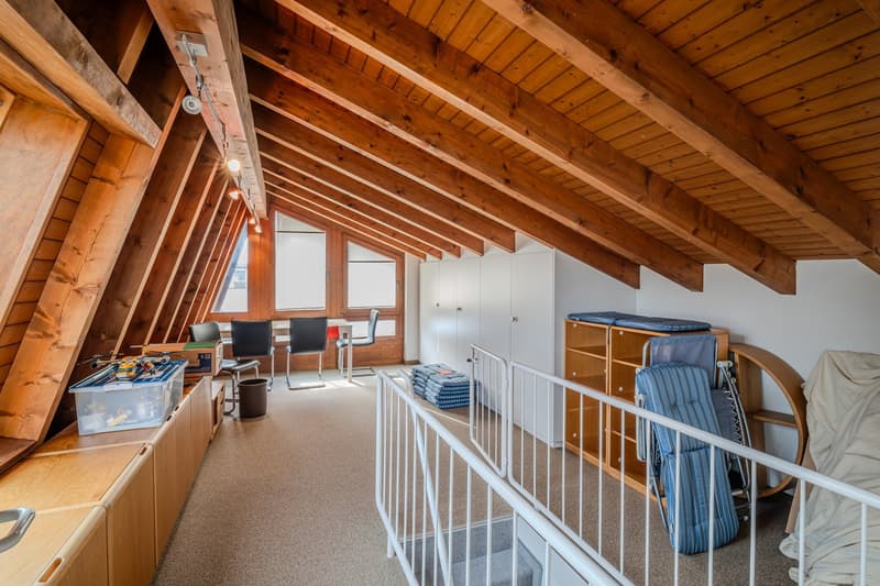 attic room with wooden beams, carpeted floor, white railings, wooden cabinets, white wardrobe, desk, chairs, storage boxes, and a trash bin