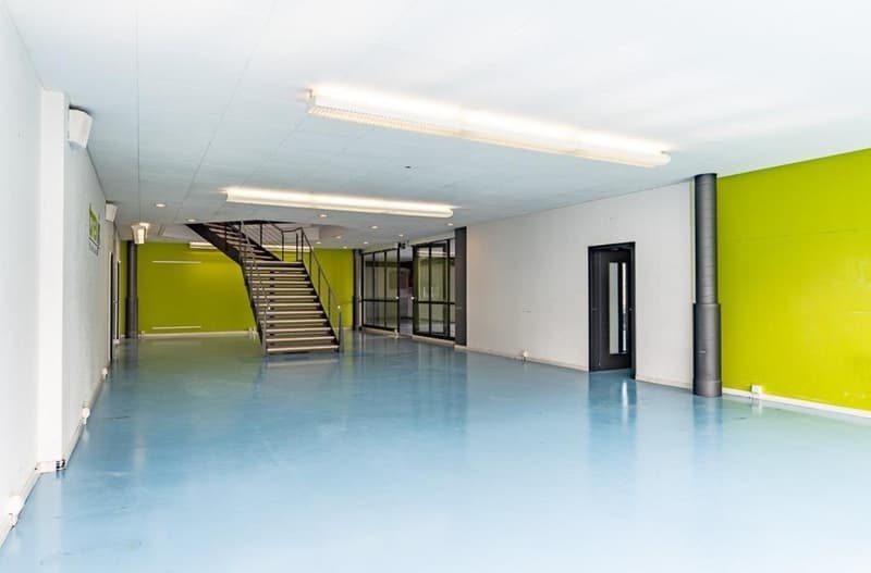 Large open space with concrete flooring, white walls, and green accent walls. There is a metal staircase leading to an upper level. The space appears to be a commercial or office area with fluorescent lighting fixtures.