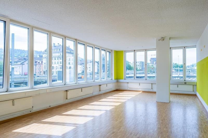 Spacious apartment with large windows offering a scenic view of the city. The room has hardwood floors, white walls, and a bright yellow accent wall. The apartment appears to be in a high-rise building with a modern, open layout.