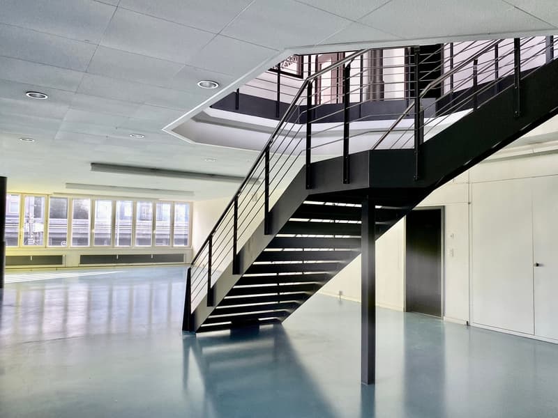 The image shows a modern, open office space with a large staircase in the center. The space has high ceilings, large windows, and a sleek, minimalist design. The staircase features a black metal frame and glass railings, creating a visually striking focal
