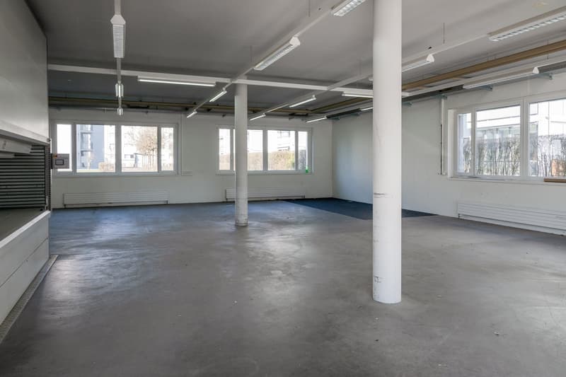 Open floor with concrete flooring, large windows, central support pillar, ceiling lights, white walls, industrial ventilation units, drain in the floor