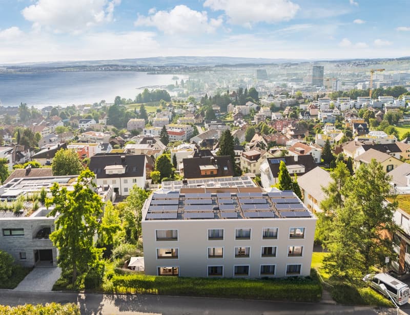 Modern apartment building with solar panels on the roof, surrounded by trees and greenery, with a view of the city and the lake