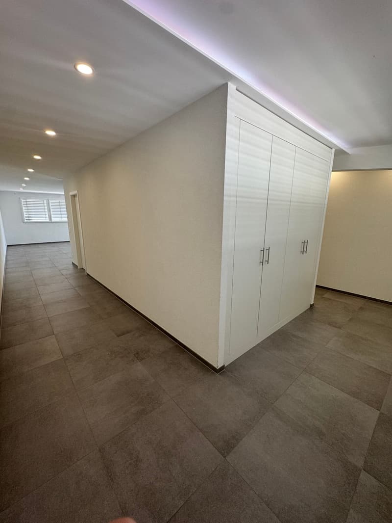 The image shows a spacious room with white walls, a tiled floor, and built-in white cabinets along one wall. The room appears to be well-lit with recessed lighting in the ceiling.