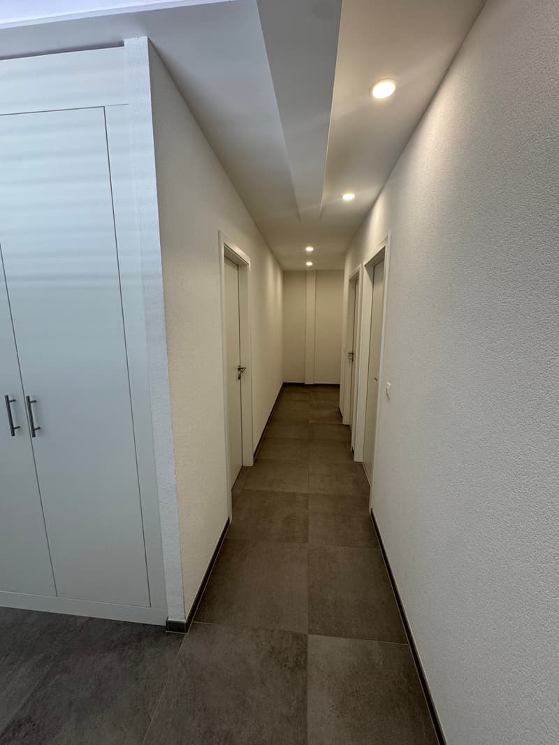 The image shows a long, narrow hallway with a tile floor and white walls. There are several doors leading off the hallway, and recessed lighting fixtures in the ceiling.