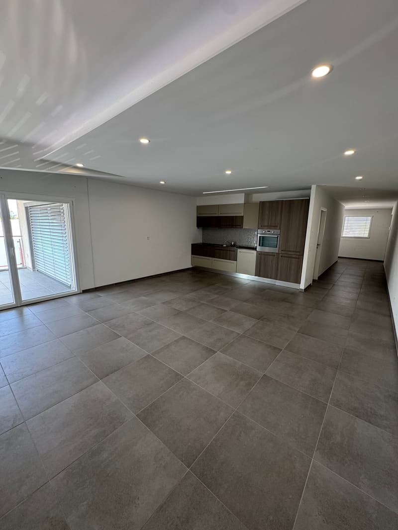 Spacious open-plan living area with large windows, grey tile flooring, modern kitchen with dark wood cabinets and a dishwasher, and recessed lighting throughout the space.