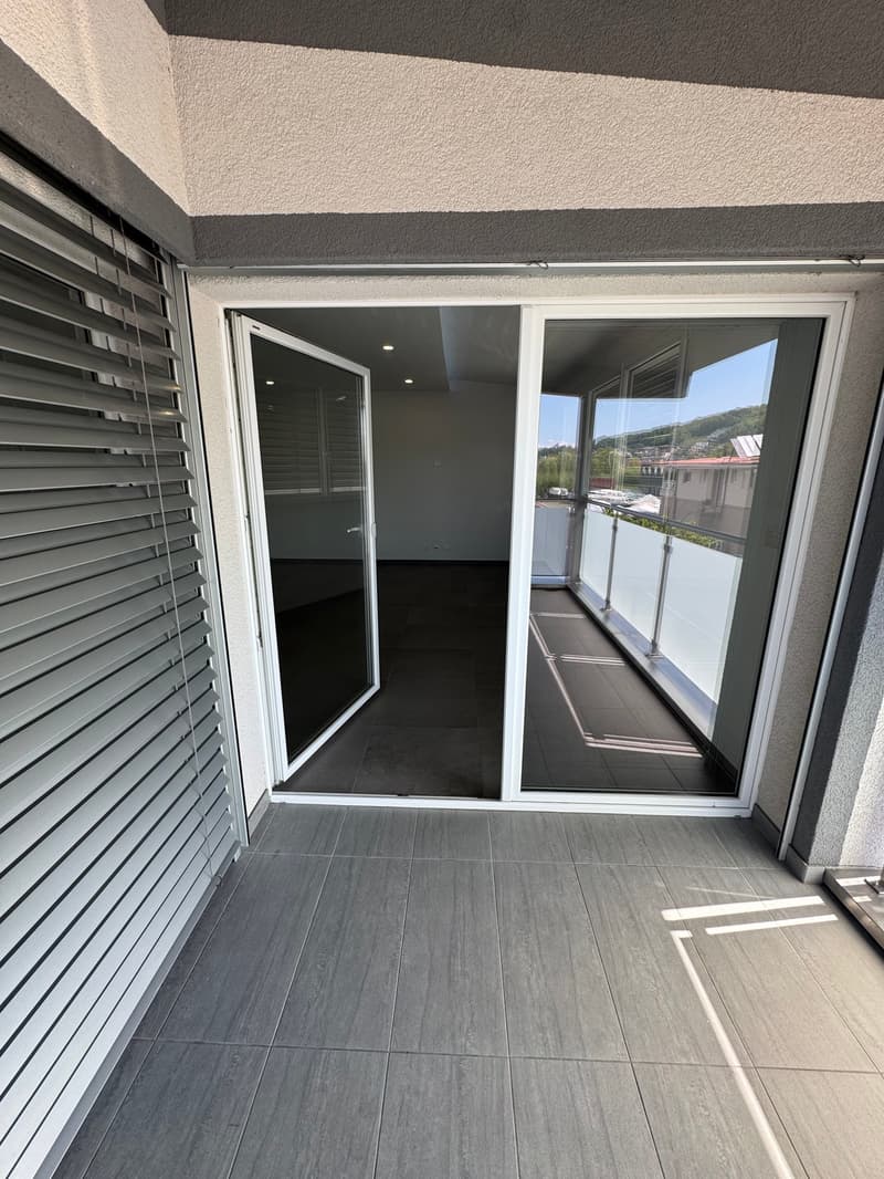 The image shows a balcony or terrace area of an apartment. The floor is covered with gray tile, and there are large sliding glass doors that lead out to the balcony. The walls are painted in a light color, and there are blinds on the windows. The balcony