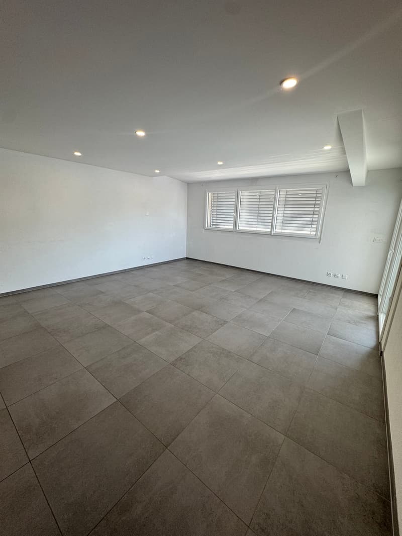 Large open room with gray tile flooring, white walls, and recessed lighting. The room has a clean, modern aesthetic.