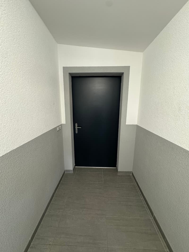 The image shows a hallway with a black door at the end. The walls are painted in a light color, and the floor is covered with gray tiles.
