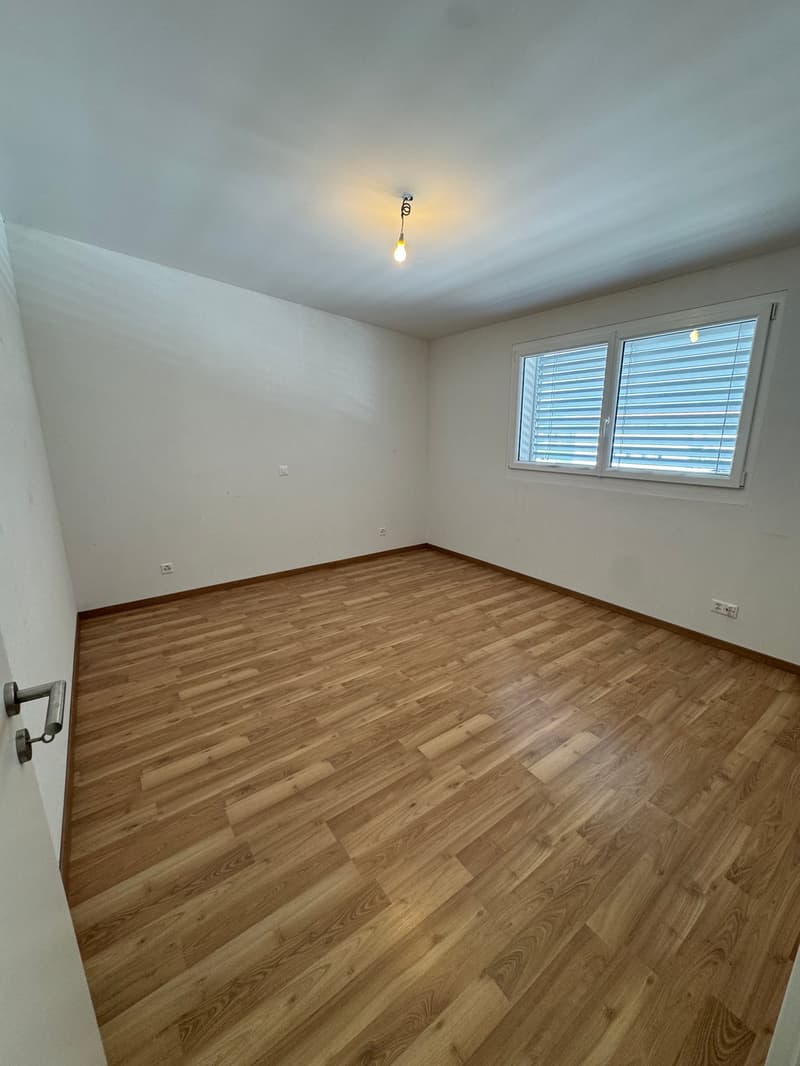 This image shows an empty room with white walls and a hardwood floor. The room has a single window with blinds, and a light fixture on the ceiling.
