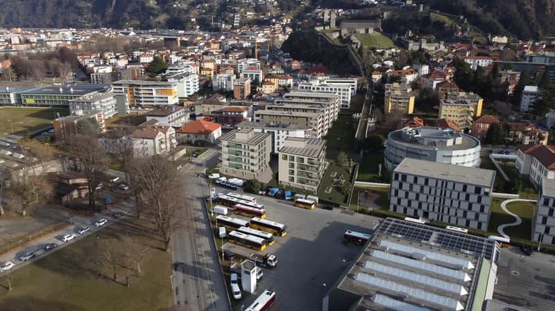 Aerial view of multiple buildings, some have solar panels on the roofs, buses parked in front of buildings, trees, parked cars and wide open space