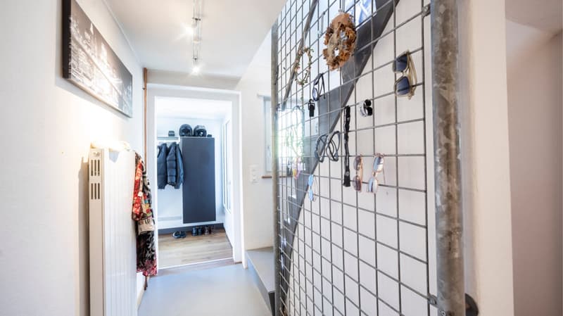 Corridor with a metal grid wall, wooden floor, white walls, a radiator, a coat rack, a cabinet, and various items hanging on the grid.
