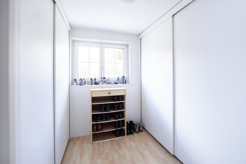 Empty room with white walls, wooden floor, shoe rack, window with sill, and ceiling light.