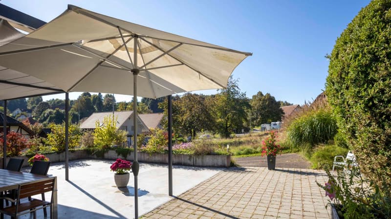 Outdoor dining area with table and chairs, large umbrella, flower pots, brick pathway, greenery, distant houses, and trees