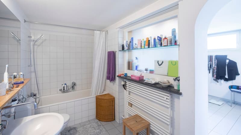 Bathroom with bathtub, shower, sink, toilet, shelves, wicker basket, radiator, rug, and towels.
