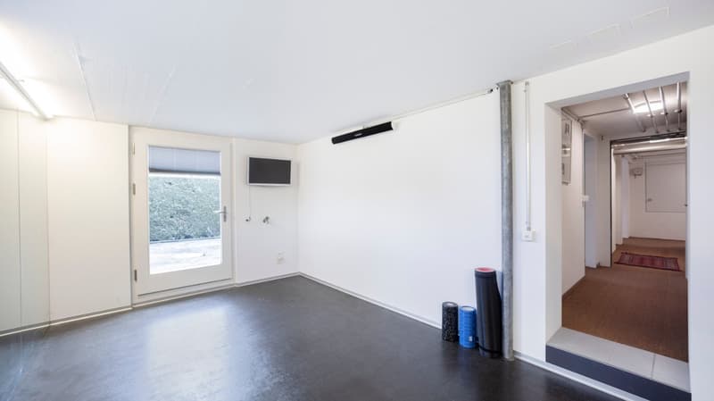 Empty room with white walls, black floor, glass door, window with blinds, mounted TV, power outlet, yoga mats