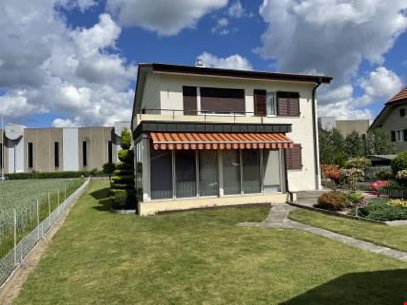 2-story house with a balcony, garage, and parking space. The house has a brown roof, beige exterior walls, and large windows. The front yard is well-maintained with a grassy lawn and landscaping.