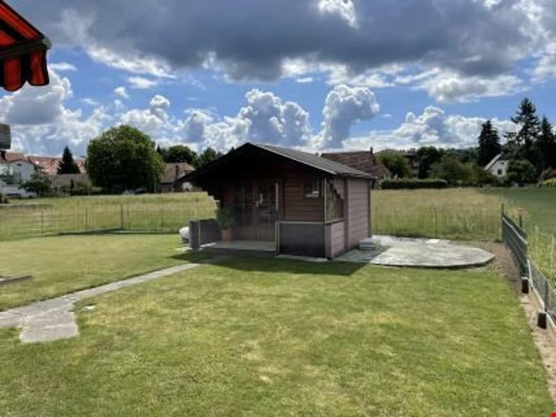 Single-story wooden house with a garage, surrounded by a grassy yard and trees. The sky is partly cloudy with some dramatic clouds.