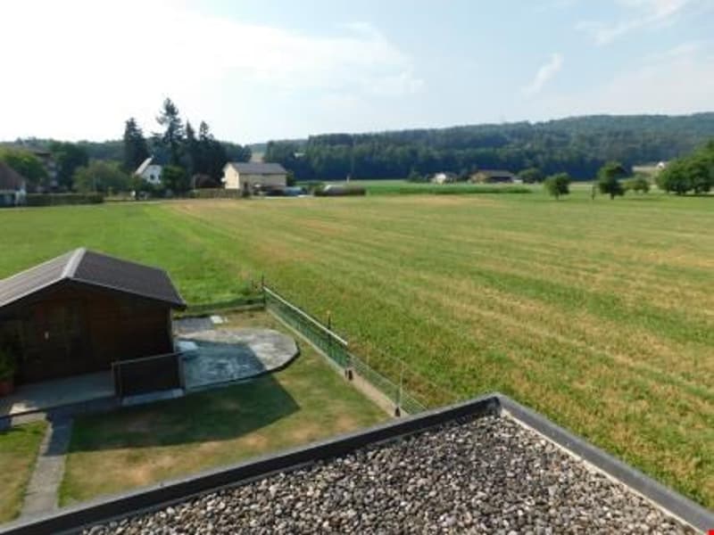 Scenic rural landscape with a large open field, trees, and a small building or shed in the foreground. The image appears to be taken from a balcony or terrace overlooking the countryside.