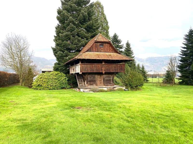 Old wooden house with a red roof and a balcony on the upper floor, surrounded by a green lawn and trees.