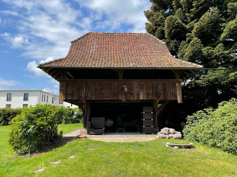 Traditional barn-like building, red tiled roof, wooden walls, open entrance, garden setting, modern building in the background.