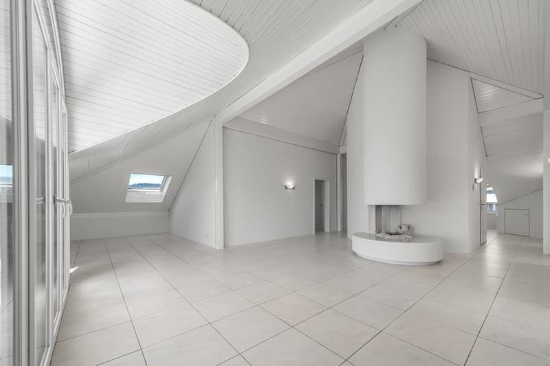 Spacious attic or loft space with high ceilings, white walls and floors, a round fireplace, and a skylight window providing natural light.