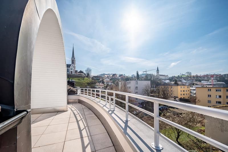 Balcony with a view of a town, including a church steeple, residential buildings, and a sunny sky with some clouds.