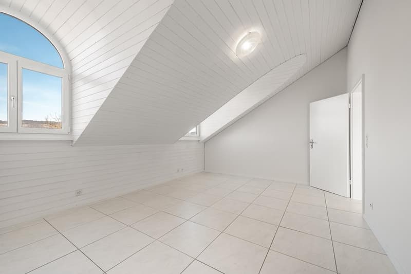 Spacious attic room with white walls, white paneled ceiling, and large window providing natural light. The room has a tiled floor and a door leading to another area.