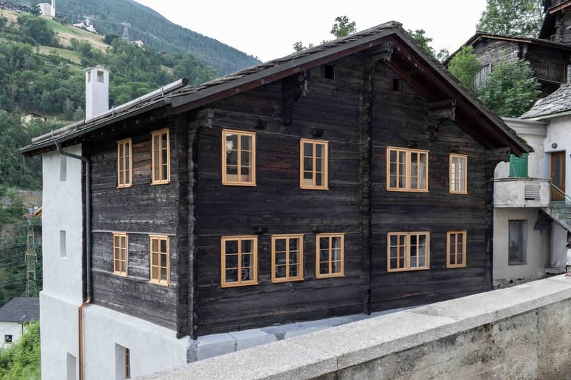 2-story wooden chalet-style house with dark wooden exterior, multiple windows with wooden frames, and a balcony or terrace visible on the upper level. The house is situated in a mountainous, forested area with other buildings and structures visible in the