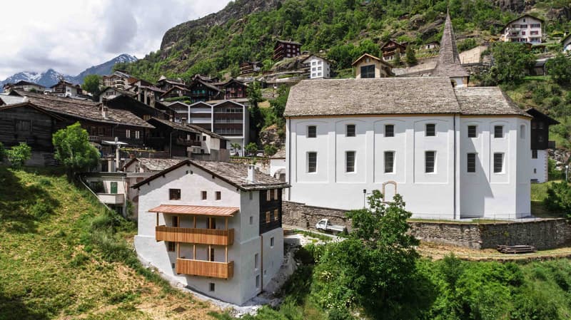 Modern house with balconies on a hillside, white exterior, stone foundation, surrounding houses, greenery, mountains in the background.