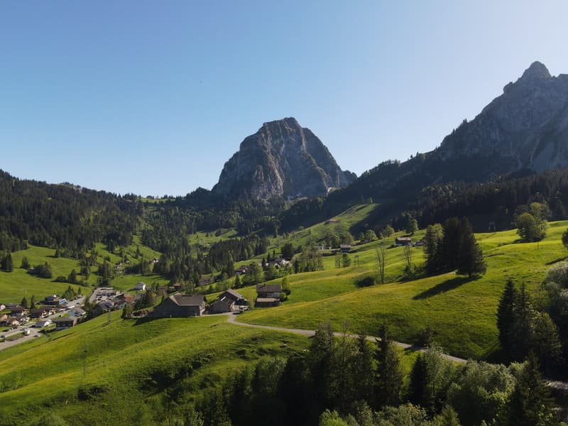 The image shows a picturesque mountain landscape with a small village nestled in the valley below. The scene features lush green meadows, dense forests, and towering rocky peaks in the background. The village appears to be composed of traditional alpine-s