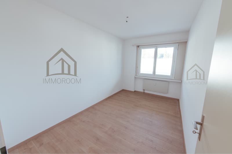 An empty room with white walls, wooden floor, and a window on the right side. A radiator is attached to the wall near the window.