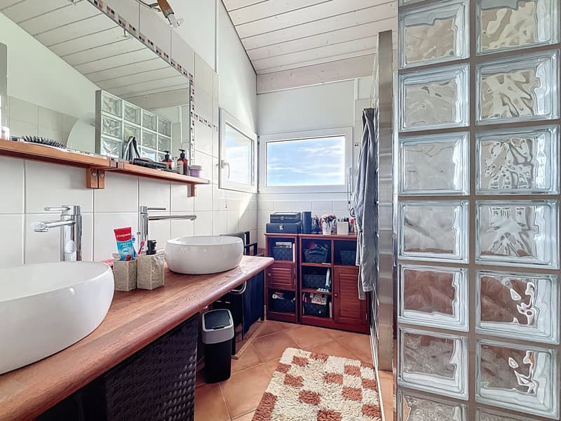 modern bathroom with double sink, mirror, wooden shelves, tiled floor, glass block wall, wooden cabinet, rug