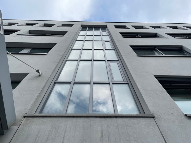 The image shows the exterior of a modern, multi-story building with a distinctive architectural design. The building features a large, vertical window panel that spans multiple floors, surrounded by a grid-like pattern of windows and concrete walls. The o