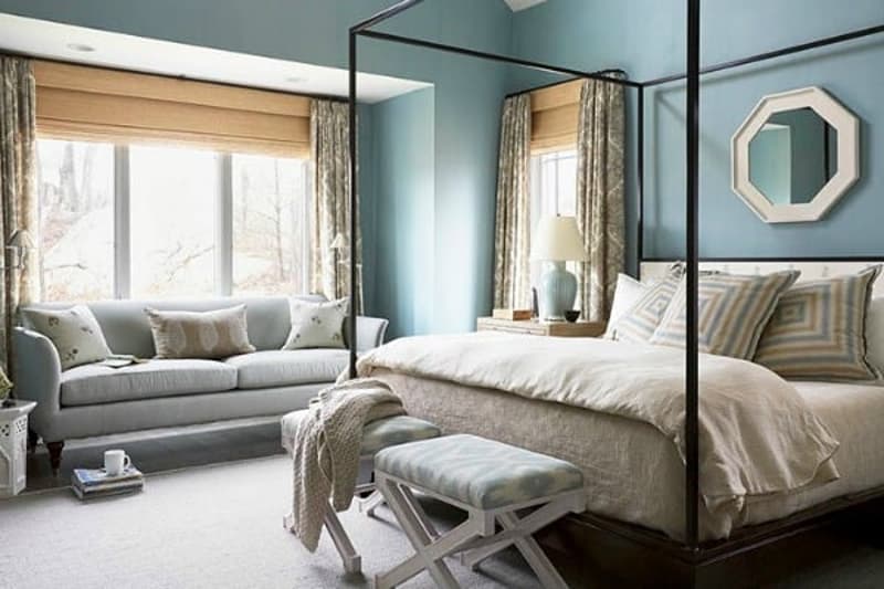 Spacious bedroom with a four-poster bed, large windows, gray and beige color scheme, sofa, and an octagonal mirror on the wall.