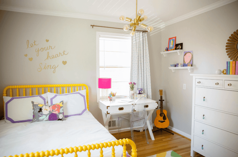 Bright and colorful bedroom with yellow metal bed frame, floral patterned bedding, white dresser, guitar, and a chandelier. The room has a cozy and creative vibe with wall decor and shelves displaying personal items.