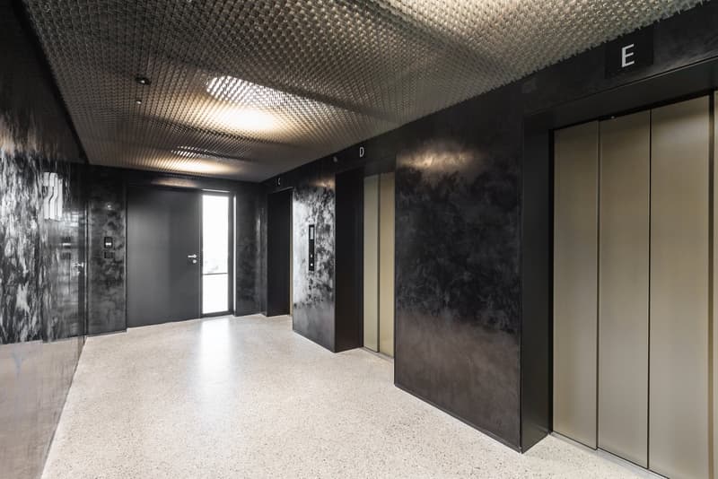 An empty hallway with shiny, patterned walls and shiny floors, four elevator doors labeled E, D, C, and 22