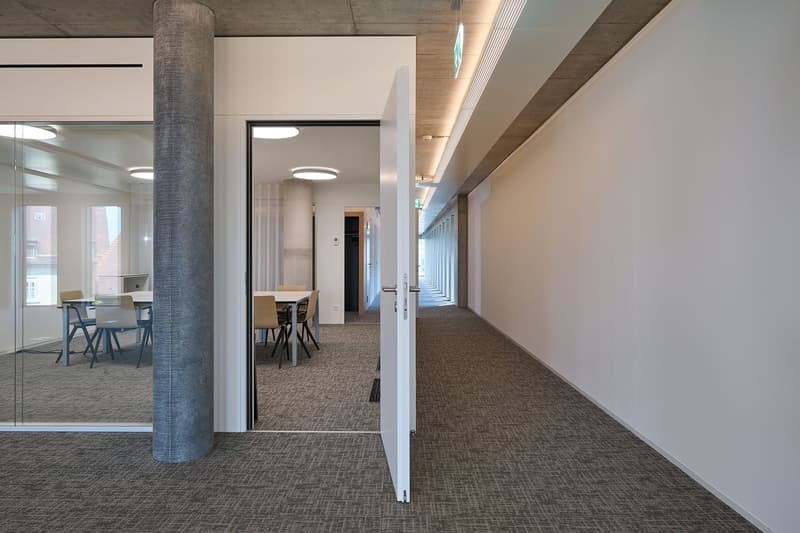 Empty room, carpeted floor, open doorways, tables, chairs, ceiling lights, glass walls.