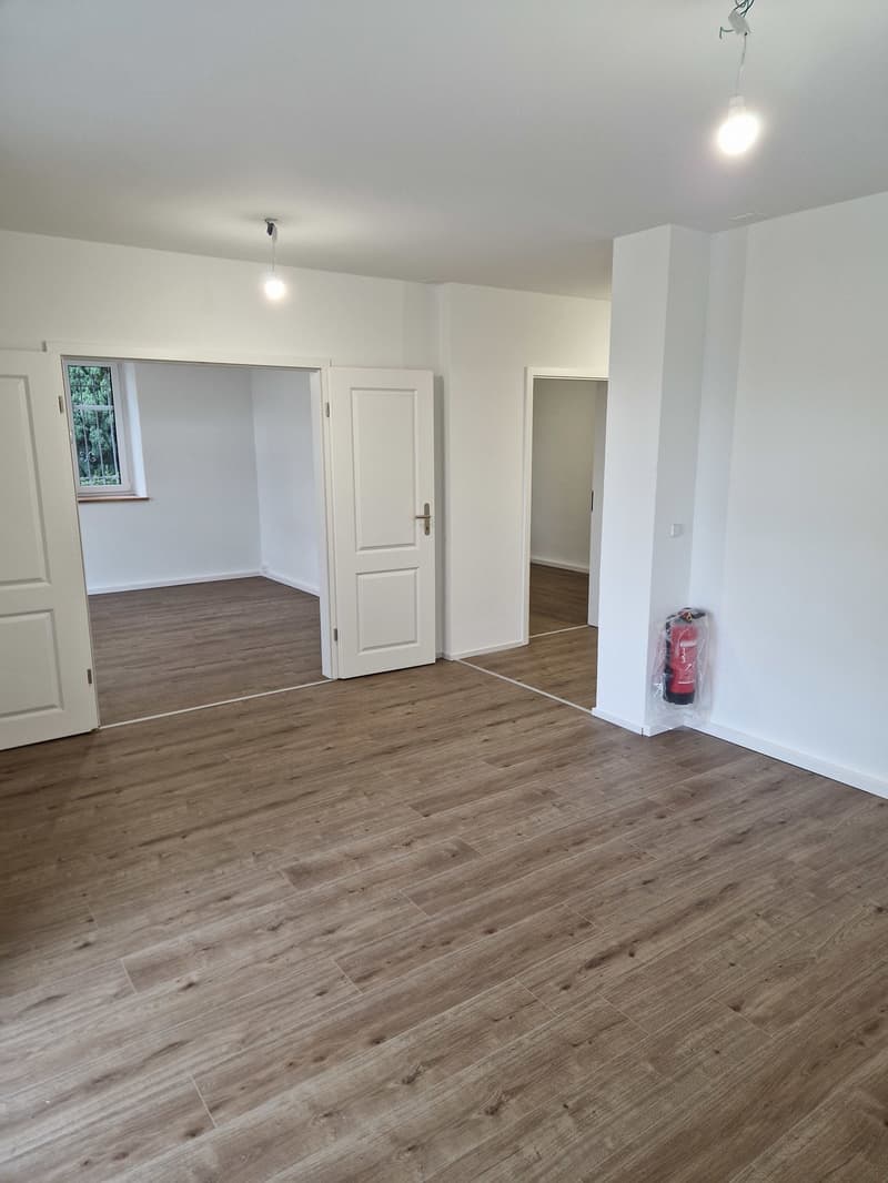 Empty room with wooden floors, white walls, two doors, and a window with a view of the outside.