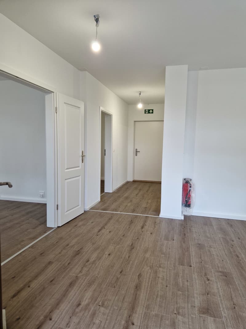 Empty room with wooden floor, white walls, two doors, and a fire extinguisher