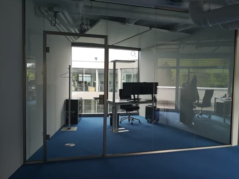 An empty office with glass walls, a desk with monitors, and a blue carpet.