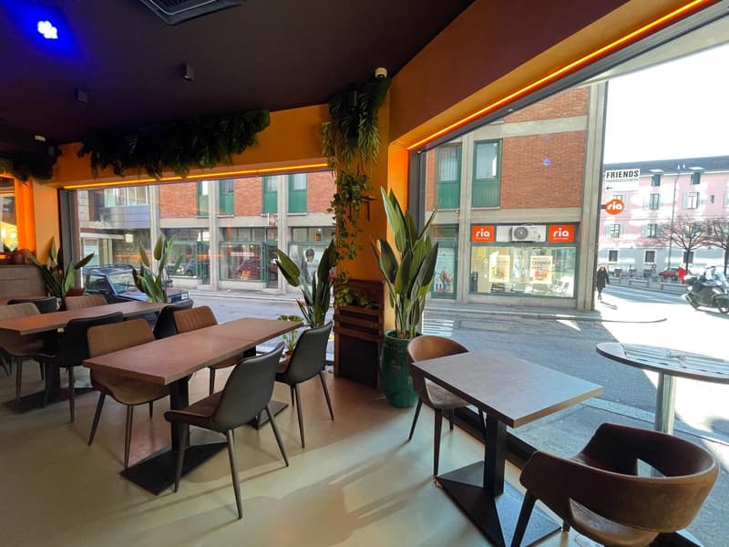 A restaurant with tables and chairs, wooden tables, plants, and a view of the street outside.