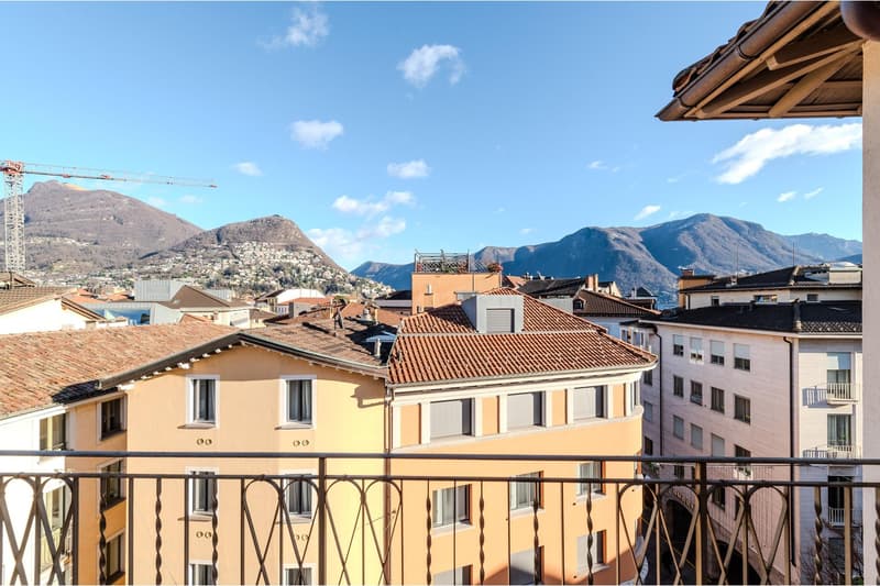 Building with multiple floors, red roof tiles, balcony, mountains in the background