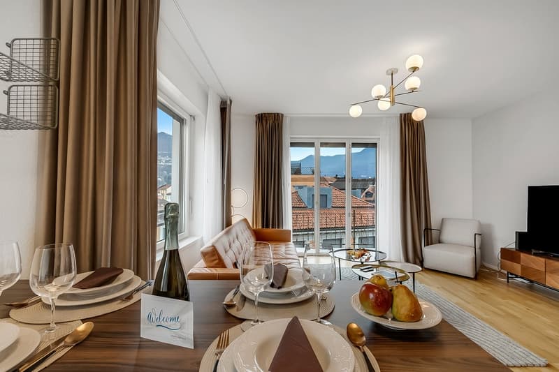 dining room with a table, wine glasses, a bottle, a welcome card, a couch, a TV, a lamp, a rug, and a window with a view